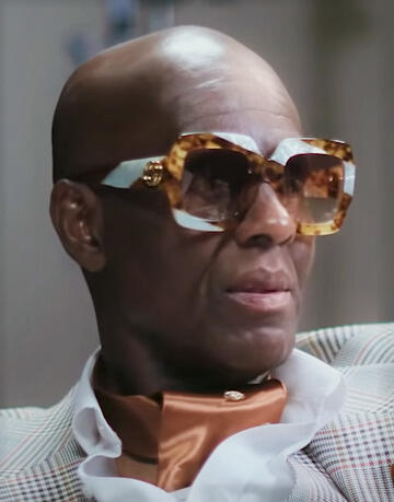 A portrait of a man wearing large sunglasses, a cravat, and a dress shirt. 