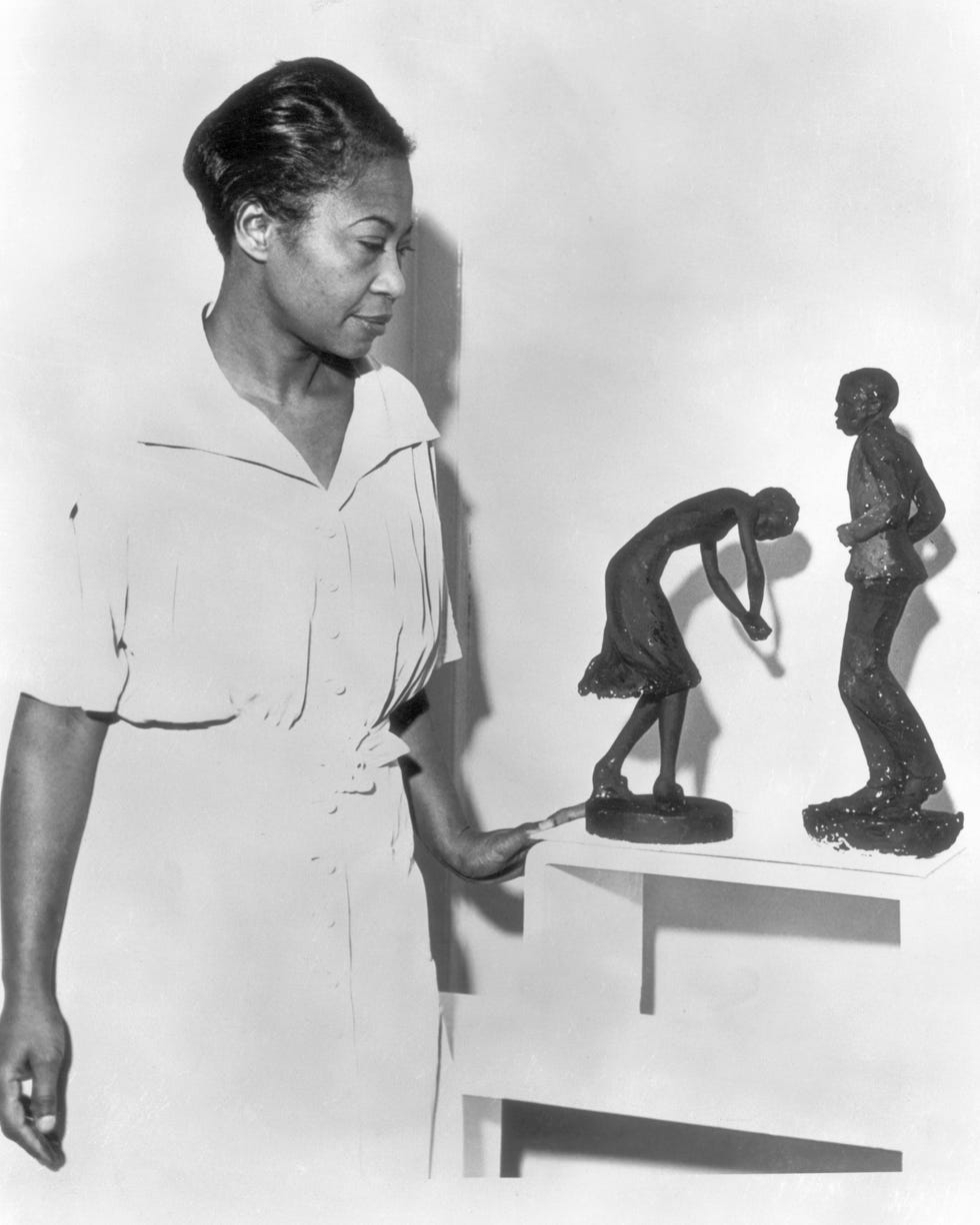 augusta savage resting her left hand on a table as she looks at two of her sculptures