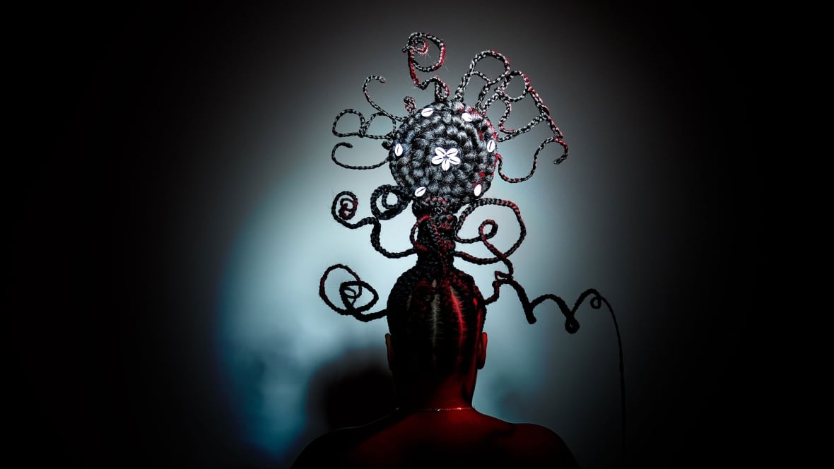 A Black woman is photographed in a medium shot, and captured with her back to the viewer. She is illuminated by a spotlight, surrounded by darkness. Her hair has been braided with wire to form a towering sculpture. The focal point is a round twist of coild braids that has been embellished with white shells. On top of the disc, braids spell "CBC Arts" in all caps.