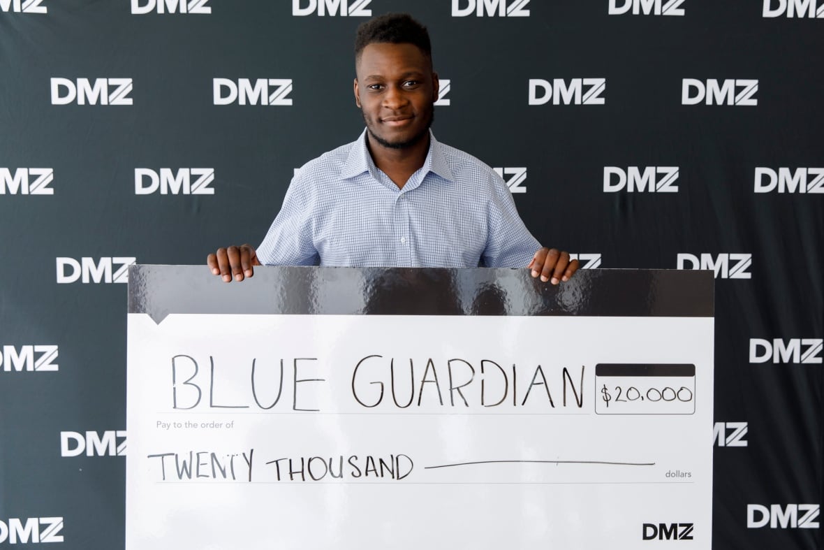 A tall Black man stands in front of banner that says DMZ, holding up a giant cheque for $20 000. He won the money for his business. 