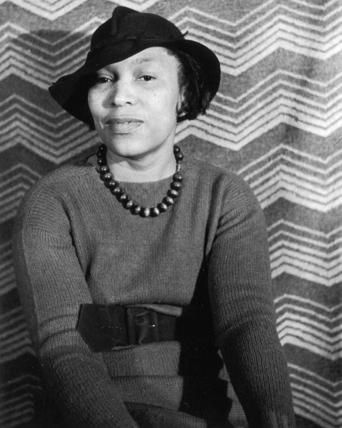 zora neale hurston wearing a sweater and hat and sitting for a photo in front of a striped backdrop