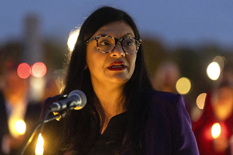 A woman wearing glasses with long black hair stands behind a microphone